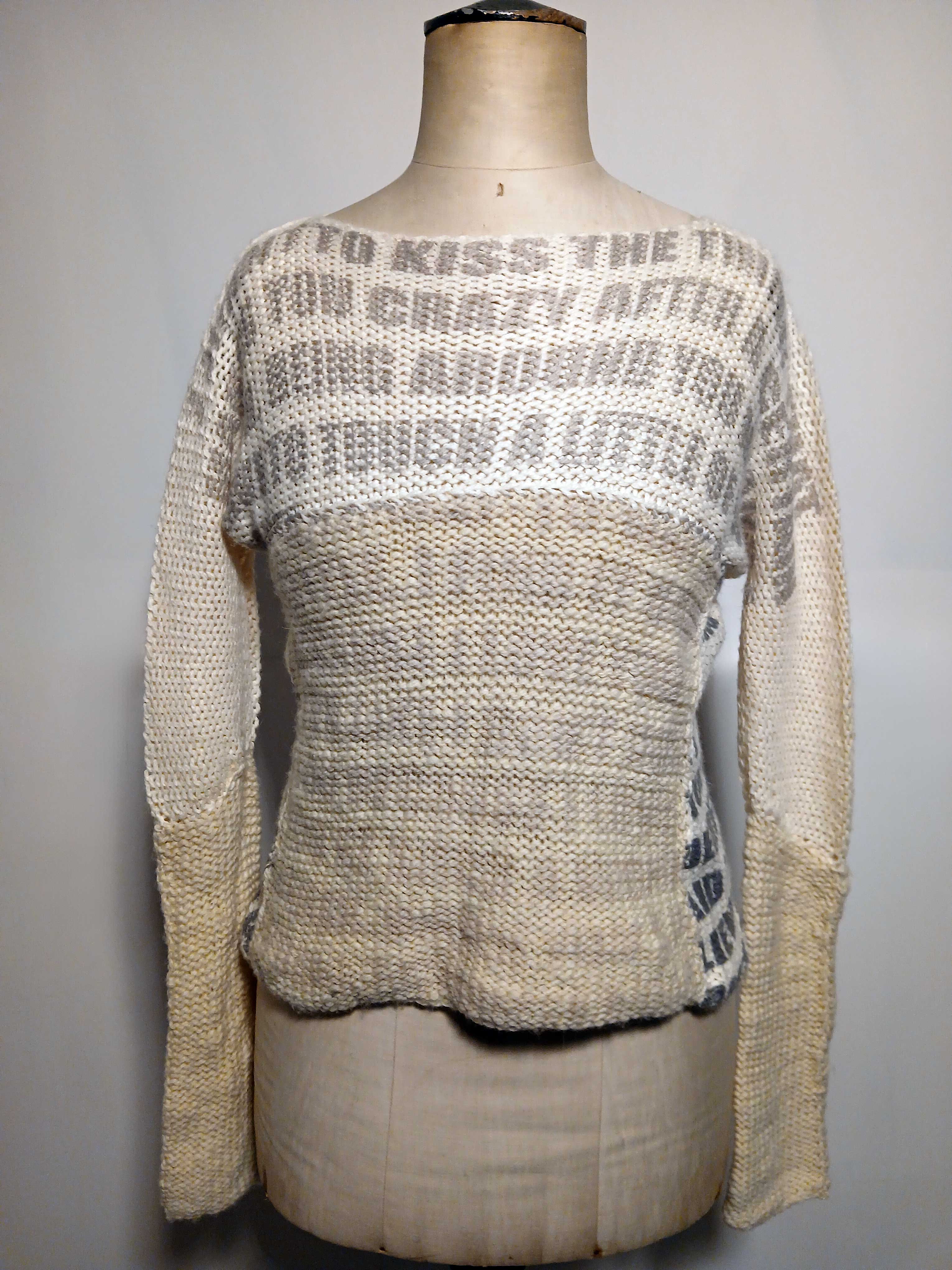 An image of a dress form wearing a sweater. The sweater is half white half dirty cream colored and has faded text written on the top and side of it. It has very faint text written on the middle. The text partially reads 'to kiss the' and 'crazy after'.