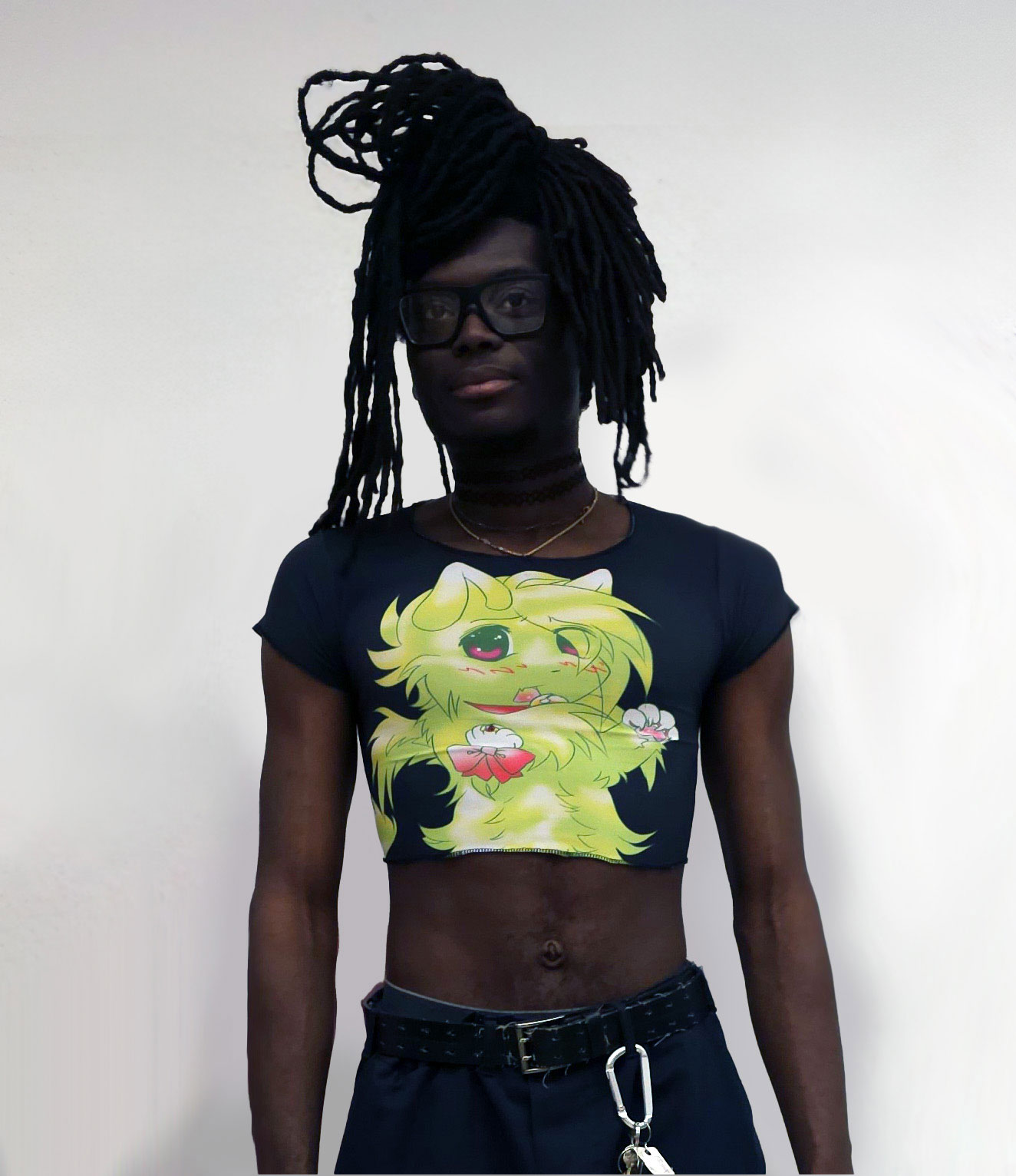 A picture of a black man wearing a cropped black shirt with a drawing of a green cat on it. The cat seems upset and has a hot pink collar with bell and a hot pink bow tied to its arm.