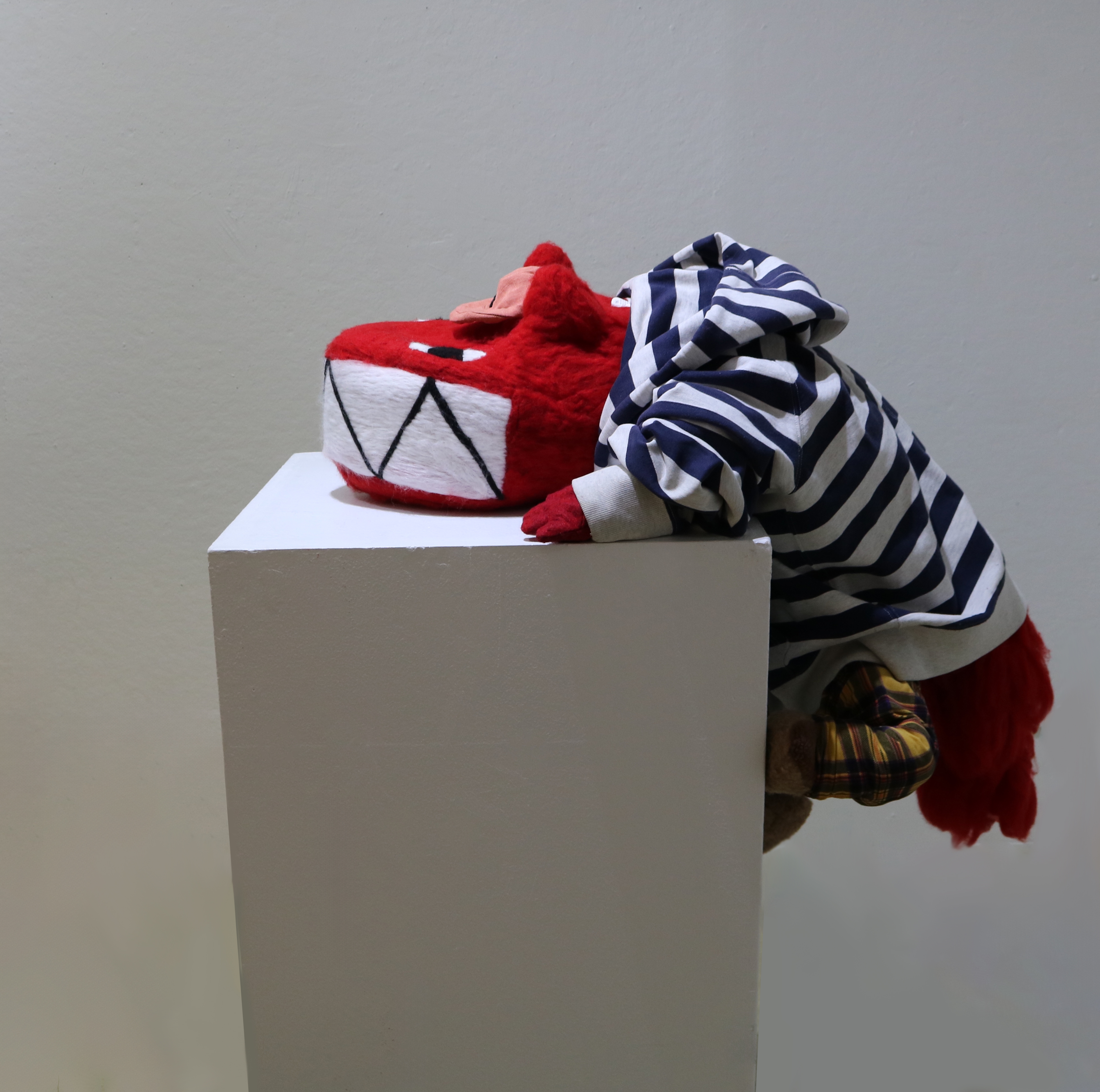 An image of a sculpture that is made of felt and clothes. It is a red fox-looking creature with a big head and a big rectangular mouth full of sharp teeth. It is hanging off the edge of a pedestal, seemingly trying to hang on to it. It has rounded cone like ears and no nose. It has a pink cap and is wearing a striped black and white hoodie. It is wearing plad yellow pants and brown shoes. It has a big bushy tail.