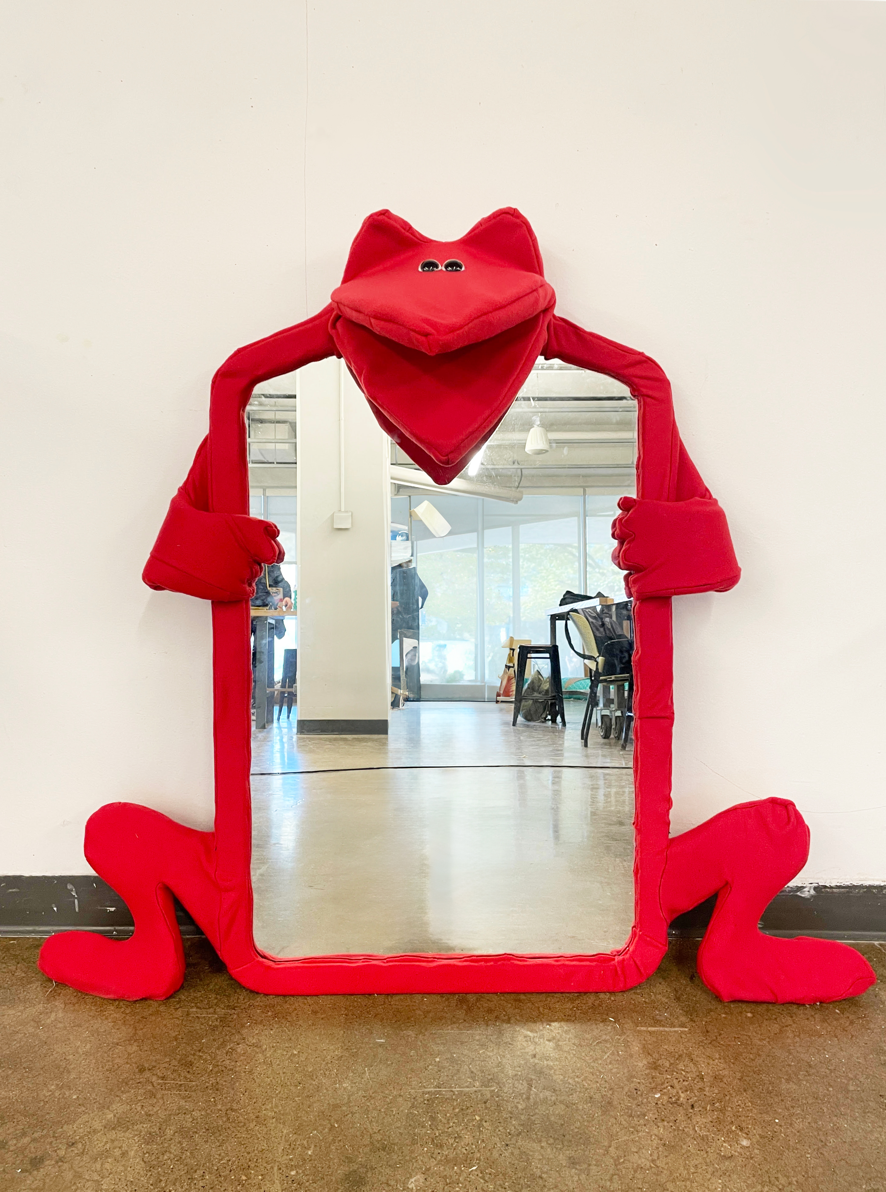 An image of a sculpture made of a repurposed mirror. It is a mirror with an fox looking creature's head, two arms and two legs sticking out of it. The creature looks happy. It has two plush eyes that are very close to eachother. It has no nose.