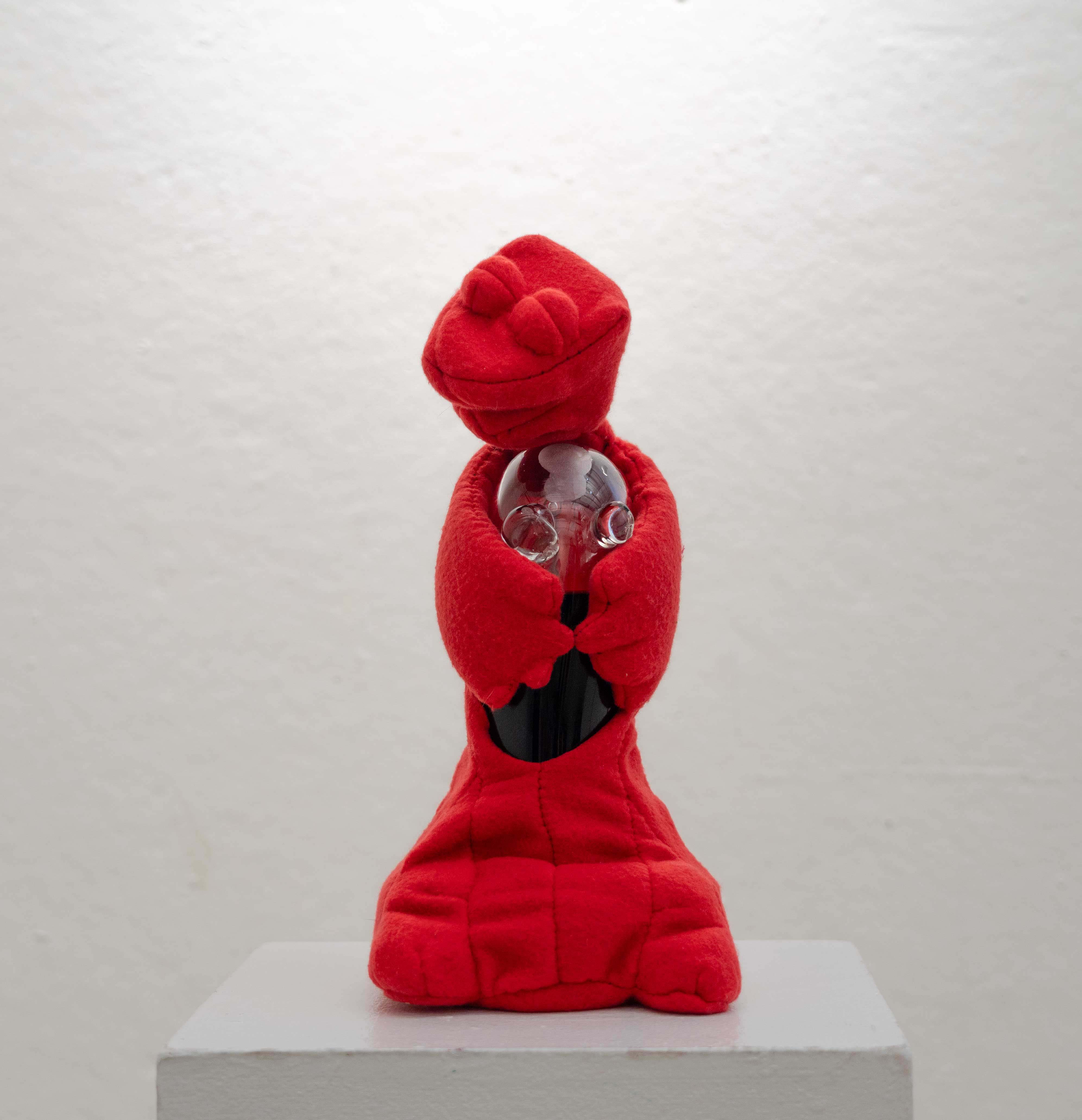 An image of a fleece soft sculpture. It is a red creature hugging a glass container with brown liquid in it. The glass container is also part of its body. The glass container is asymmetrical and has two protrusions on its top that look like nipples. The creature has a flat base with two small round feet. Its eyes are closed and it looks delighted.